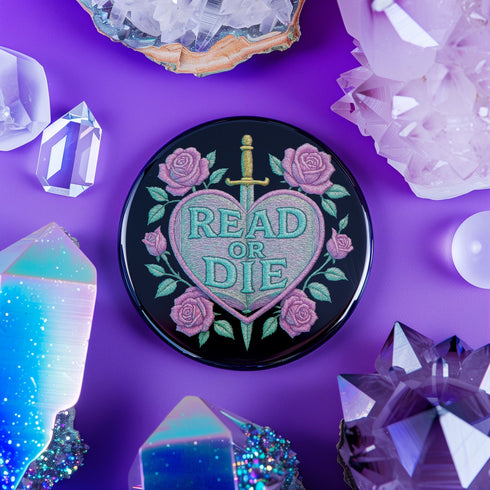Glossy black pinback button featuring embroidered-style artwork of a pastel purple heart that reads “Read or Die” pierced by a sword and surrounded by roses; shown on books, graffiti, and lace backgrounds.